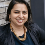 Podcasting instructor Golda Arthur smiling to camera wearing black jacket with wooden necklace and hoop earrings