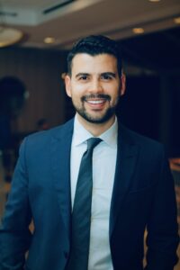 Mohamad, alumni award winner, smiles to camera wearing a dark blue suit with tie. He has dark hair and a beard and moustache.