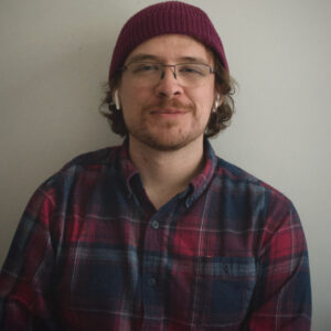Alan is light skinned wearing a wool hat, wearing glasses and his earbuds, and plaid shirt.