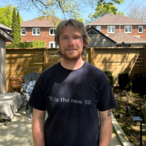 Connor stands in a backyard shaded by an unseen tree with a fence and two houses visible in the background.