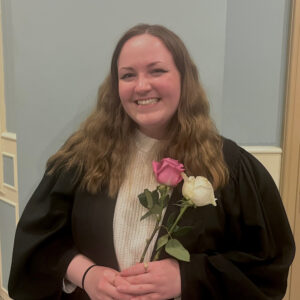 Erin is light skinned female with long, light brown hair. She is wearing a black robe over a cream sweater and she is holding a pink and a white rose.
