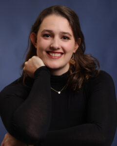 Karina is light skinned with long dark hair. She wears a black sweater and is smiling to camera.