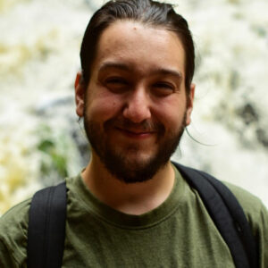 Marcus has dark hair and a beard. They are wearing an olive green tshirt and backpack straps are visible. They are outside with greenery in the background.