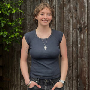 Grad Sam Flood stands outside in front of a wooden fence with vertical slats. She is light skinned with short light brown wavy hair.