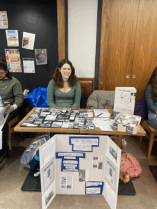 a woman sits behind a table at a fair with photos displayed on the table and board display of yearbook society