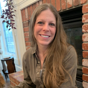 Tamara is light skinned with long, light brown straight hair. She sits on a hearth in front of a red brick fireplace.