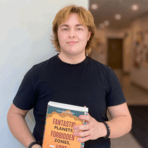 Bailey stands against white wall wearing black tshirt - holding a book