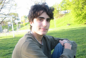 Oliver sits on the grass in a park with trees and a soccer pitch in the background.