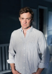 Patrick wears an open necked casual shirt and is standing outside a building lit by sunshine