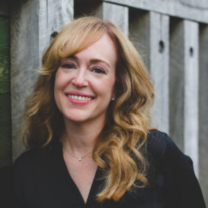 Jenn leans against a wooden fence. She has long, hair and is wearing a dark shirt.