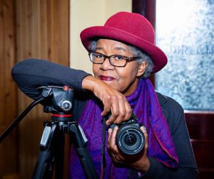 Book launch Tender by Sylvia Hamilton - here in hat and scarf holding a camera and leaning on tripod