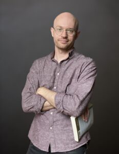 Mike stands casually with arms crossed a book in his right hand, against neutral gray background. He is a light skinned male, bald with glasses. He wears an open necked shirt