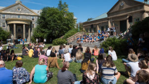 Wide shot of Quad looking toward Alex Hall as students sit on grass and listen to the welcoming ceremony on the Library steps.