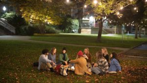 A group of eight students sit together on a blanket on the quad at night.