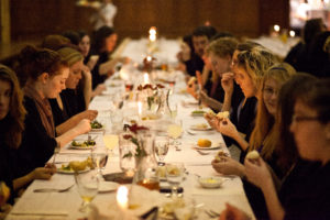 Students in academic gowns sit along two sides of a formally set table eating a meal.