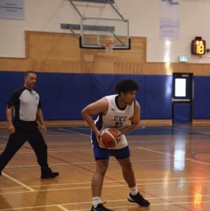 Caleb Rennie on the basketball court, holding a ball, with referee behind watching the play