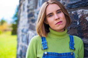 Hermione Davis leaning left shoulder on large stone wall with green lawn in background