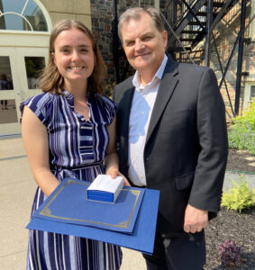 A young woman with shoulder-length brown hair in a blue striped dress poses next to an older man. She is holding an award. They are outside in a garden.