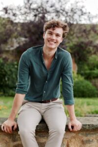 Nick Harris, sits on a stone wall with trees, grass and shrubs behind him. He wears a green shirt and taupe jeans.