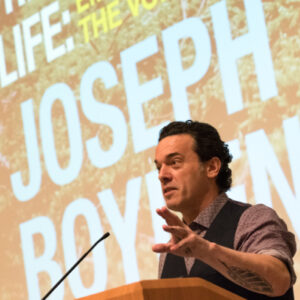 Writer Joseph Boyden speaking at podium with big image of his book cover in background