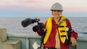 Emily wearing hard hat and safety gear and carrying an DSLR camera with boom mike attached on a boat