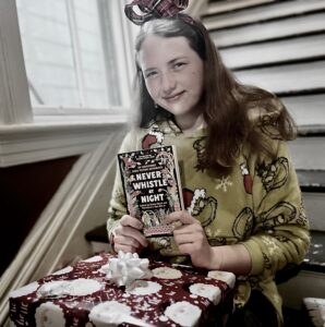 Emily sits on the stairs holding a present on her lap and a book in her hands to camera