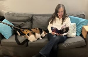 Gillian Turnbull sits on her couch reading her book with aghast look on her face - her cat sits between her and a guitar leaning on the other end of the couch