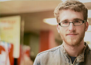 Alex Fountain in bookstore in grey denim shirt and wearing glasses looking to camera on far right of frame