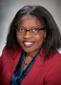 Eunice Abaga wearing red blazer and blue blouse, shoulder length hair and glasses, smiling to camera in formal portrait.