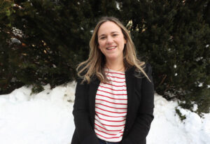 Stephanie is wearing a red and white striped shirt under a black blazer. She stands in front of a snowbank with evergreens in the background