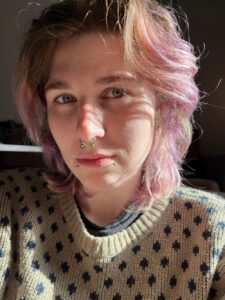 Ashton is wearing a dotted sweater and has pink highlighted hair. He sits in the sun looking to camera.