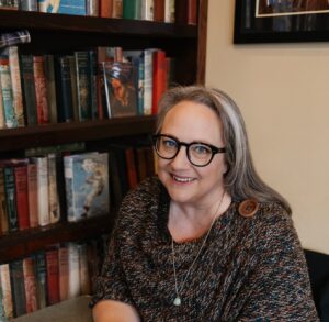 Simone sits in front of a bookcase. She wears a dark sweater, has long grey hair and black framed glasses