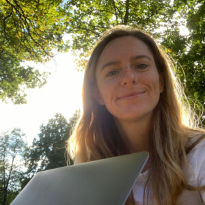 Michaela sits under a tree with the sun behind lighting up her long light brown hair, her laptop lid visible at the bottom of the shot.