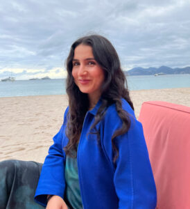 Elia has long dark hair, light skin, and is wearing a royal blue jacket and sitting outdoors on the beach in Vancouver. The north shore mountains and sea are in the background.