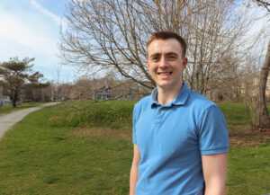 Jack is light skinned male with buzzed short reddish brown hair. He wears a light blue golf shirt and is standing outside in a park setting with early spring trees in the background.