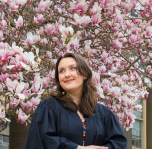 Jessica is a light-skinned female with long dark hair. She is wearing an academic gown and looking off to her right with a grin. Behind her is King's now famous magnolia.