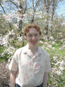 Kyle is light skinned and has short, wavy, red hair and is wearing a light flowered shirt. Kyle is standing in front of a white magnolia tree.