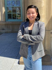 Lokman is Asian with shoulder length dark hair. She is wearing a grey suit jacket, black shirt and jeans. She stands with her arms crossed outside the windows of the King's Library on a sunny morning.