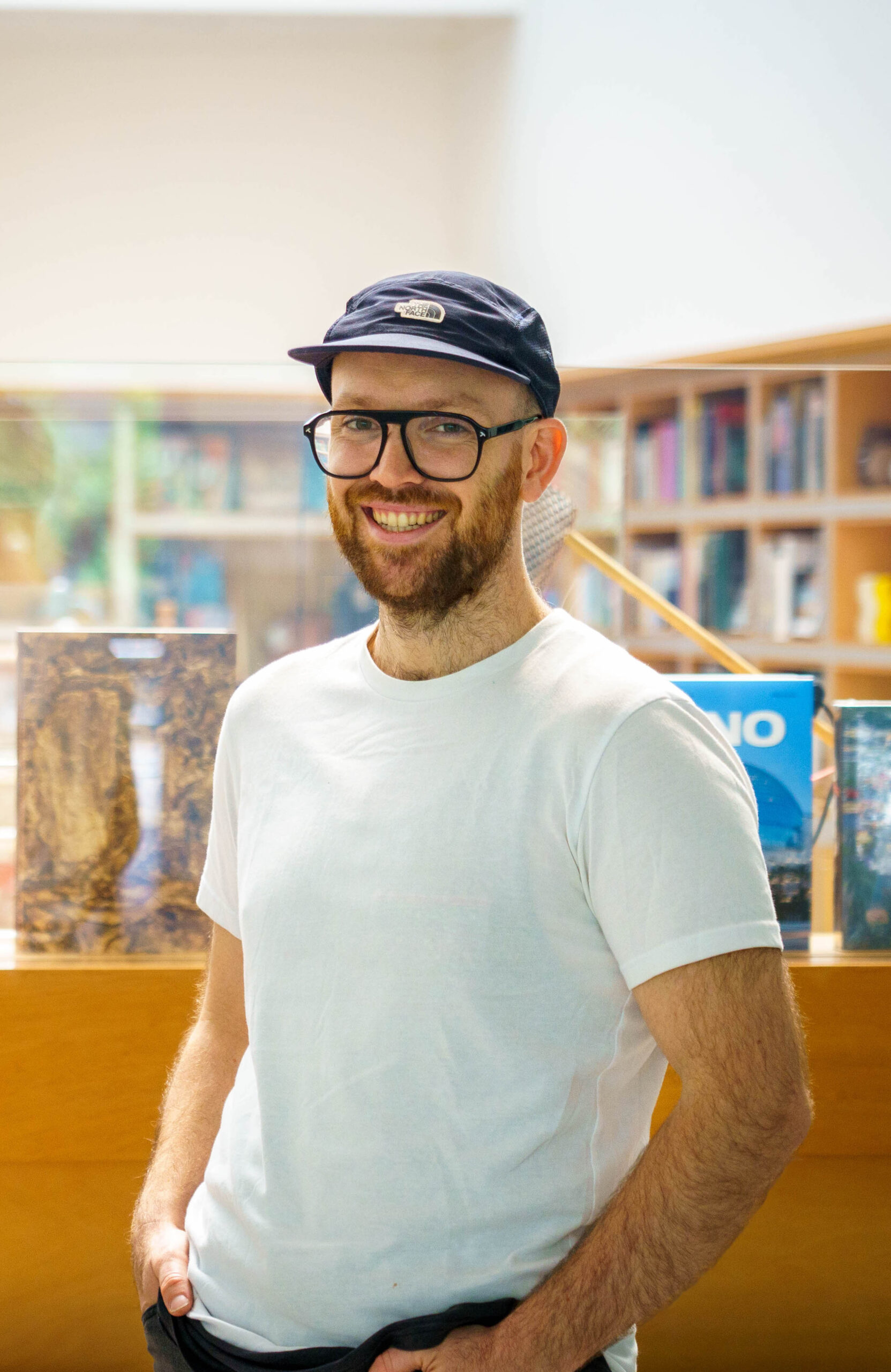 Patrick wears a blue hat with brim, large black framed glasses. He is fair skinned and has a full beard. He wears a casual white tee stand with hands in pockets with bookshelves in background.