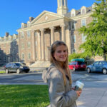 Rachel is light skinned with long blond hair. She stands sideways to camera and smiling. The King's A&A building is behind her and the sun is shinging and the sky is brilliant blue.