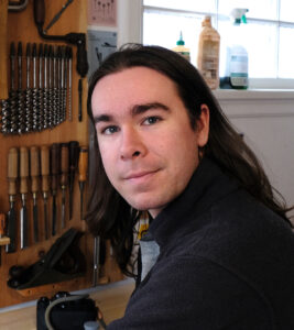 Ronan, a light-skinned male with long dark hair sits in front of a meticulous tool bench with a window over his shoulder.