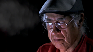 Elder Albert is wearing a flat cap and red shirt. He is looking down and light is reflected in his glasses.