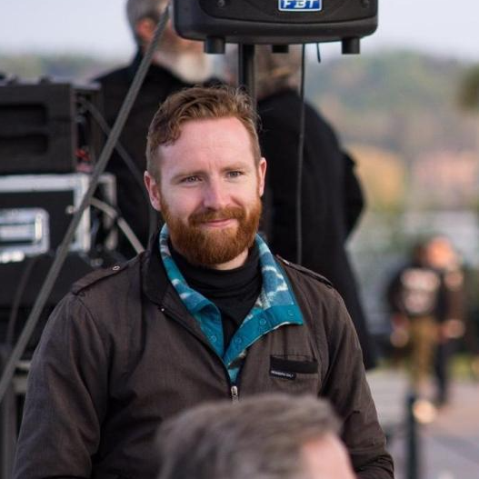 Sansom has light skin, red short hair and a ginger full beard. They wear a dark jacket with a turquoise and white patterned fleece underneath. They appear to be backstage at an outdoor theatre venue with staging equipment in background.,