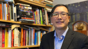 Simon stands in front of his bookcase wearing suit jacket and open necked blue shirt