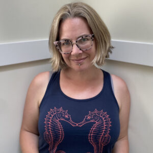 Adrienne stands in a corner wearing a blue tank top with two seahorses kissing. She is a light-skinned female with a blond bob and she is wearing glasses.