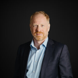 Nevin is a light skinned male with ginger hair, moustache and beard. He wears a dark suit jacket and light blue open necked shirt. He is standing against a dark background.