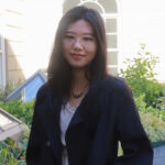 Nerissa stands outside in Willwerth Garden. She is a light skinned female with long dark hair. She wears a dark suit jacket and mauve shirt.