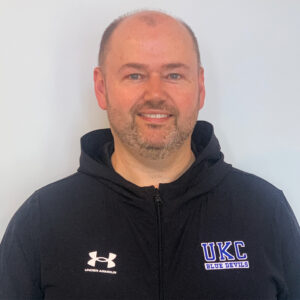 New head coach Chris Holland wears a blue UKC Blue Devils hoodie. He is a light-skinned male with a semi goatee.