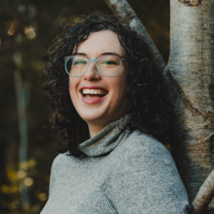 Molly stands with her back against a tree and a forest behind her. She is a light skinned female with dark, curly hair and glasses. She wears a grey sweater with a mock turtleneck.