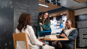 2 students sit face to face at table in Wardroom. A third stands next to a chalkboard and outlines discussion about the Odyssey.